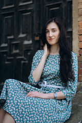Portrait of attractive young smiling woman sitting near old doors in cute summer dress with flower print