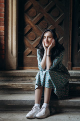 Portrait of attractive young woman sitting near old doors in cute summer dress with flower print