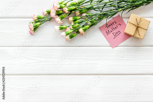 Mother's Day congratulations. Carnations and gift box on white wooden background top view copy space
