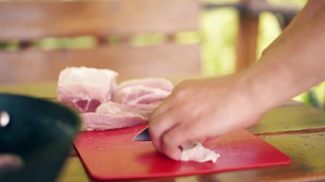 Close-up, Travelling: Young guy is standing in summer house, cooking pilaf, for picnic. He cuts meat, on red cutting board. Put it in the cauldron.