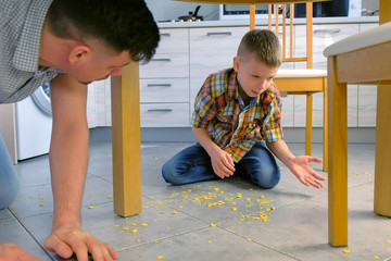 Dad scolds his son for scattered food on the kitchen floor and makes him clean up. Clean up corn...