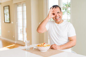 Middle age man eating rice at home doing ok gesture with hand smiling, eye looking through fingers with happy face.
