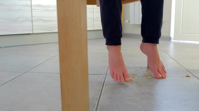 Kid eats at the table and crumbles corn flakes on the floor. Legs and dirty floor close-up.