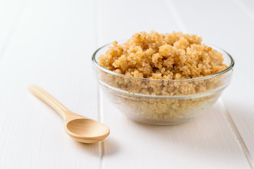 Glass bowl with boiled quinoa and wooden spoon on white table. A gluten-free boiled cereal dish.