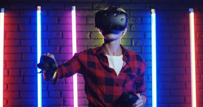 Potrait of the Caucasian blond and stylish girl in VR glasses and with joysticks playing virtual game on the colorful neon lights background.