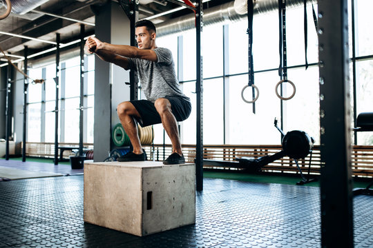 Young man dressed in black sports clothes is doing squats on the box next to the sport equipment in the gym