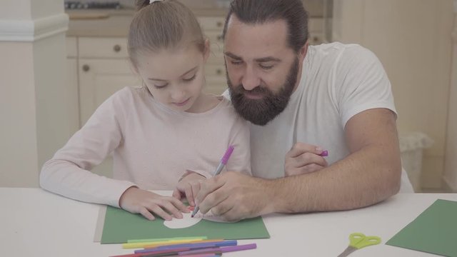 Young father and his small cute daughter are sitting in living room and drawing with markers postcard on green cardboard