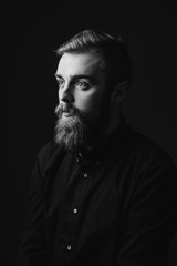 Black and white portrait of a stylish man with a beard and stylish hairdo dressed in the black shirt on the black background