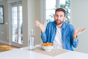 Handsome man eating pasta with meatballs and tomato sauce at home clueless and confused expression with arms and hands raised. Doubt concept.