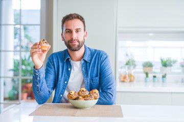 Handsome man eating chocolate chips muffin with a confident expression on smart face thinking serious
