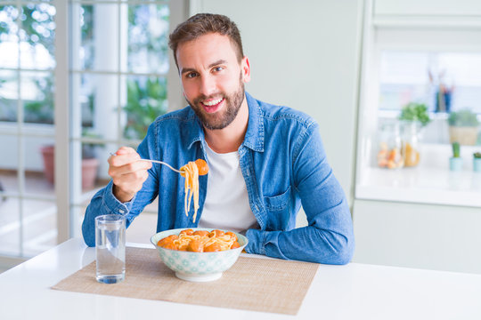 Handsome man eating pasta with meatballs and tomato sauce at home while smiling at the camera
