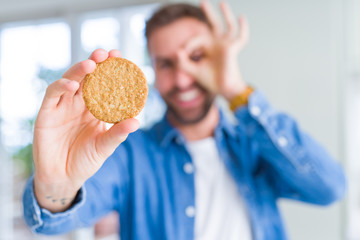 Handsome man eating healthy whole grain biscuit with happy face smiling doing ok sign with hand on eye looking through fingers