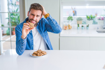 Handsome man eating chocolate chips cookies stressed with hand on head, shocked with shame and surprise face, angry and frustrated. Fear and upset for mistake.