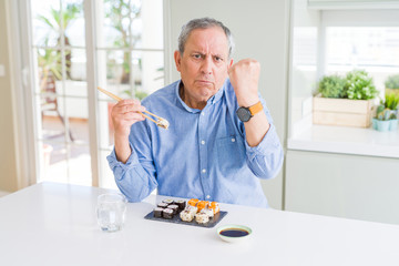 Handsome senior man eating take away sushi using chopsticks at home annoyed and frustrated shouting with anger, crazy and yelling with raised hand, anger concept