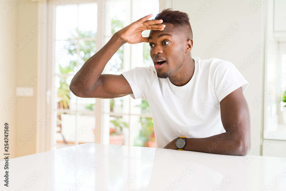 Wall mural Handsome african american man on white table at home very happy and smiling looking far away with hand over head. Searching concept.