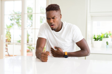 Handsome african american man on white table at home with hand on stomach because indigestion,...