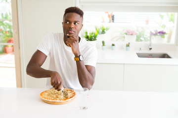 African american man eating cheese pizza at home serious face thinking about question, very...