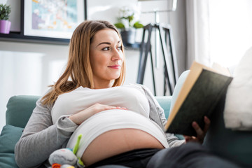 Pregnant woman relaxing on sofa reading book looking at belly