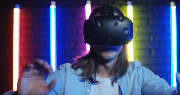 Close up of the young guy in the VR glasses having VR headset and moving like being scarry on the backgroung of neon colorful lamps.