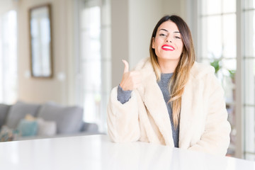 Young beautiful woman wearing winter fur coat at home doing happy thumbs up gesture with hand. Approving expression looking at the camera with showing success.