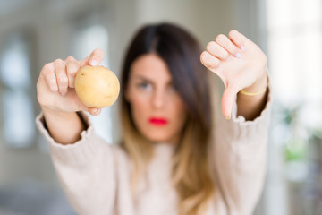 Young beautiful woman holding fresh potato at home with angry face, negative sign showing dislike with thumbs down, rejection concept