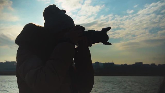 Young woman taking pictures of the sunset on the background of the sea, city and sky. Female silhouette takes pictures while holding a professional camera lens. Slow motion.