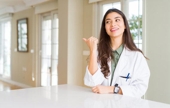 Young woman wearing medical coat at the clinic as therapist or doctor smiling with happy face looking and pointing to the side with thumb up.