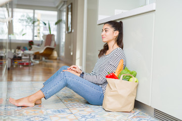 Young woman sitting on the kitchen floor with a paper bag full of fresh groceries looking to side, relax profile pose with natural face with confident smile.
