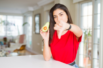 Young woman eating healthy avocado with angry face, negative sign showing dislike with thumbs down, rejection concept