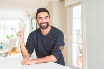 Fototapeta premium Handsome hispanic man wearing casual sweater at home smiling with happy face winking at the camera doing victory sign. Number two.