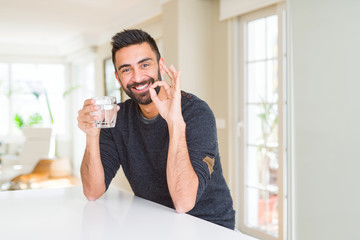 Handsome hispanic man drinking a fresh glass of water doing ok sign with fingers, excellent symbol