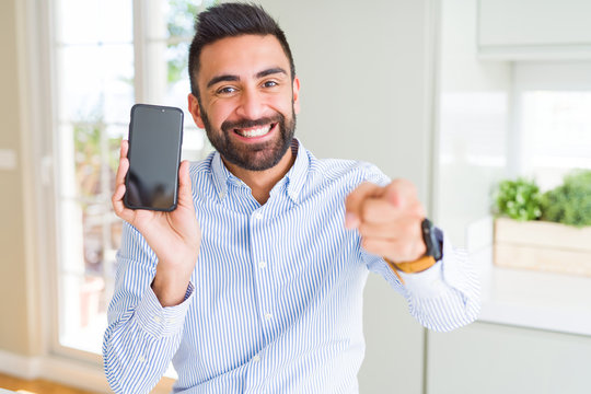 Handsome hispanic business man showing smartphone screen pointing with finger to the camera and to you, hand sign, positive and confident gesture from the front