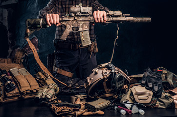 Man in a checkered shirt holding a assault rifle and showing his military uniform and equipment. Modern special forces equipment. Studio photo against a dark textured wall