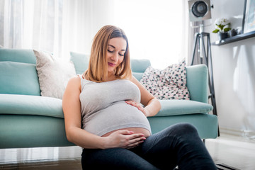 Beautiful smiling pregnant woman sitting on floor at home, touching and looking on belly