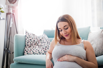 Beautiful smiling pregnant woman sitting on floor at home, touching and looking on belly