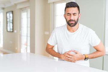 Handsome hispanic man casual white t-shirt at home with hand on stomach because indigestion, painful illness feeling unwell. Ache concept.