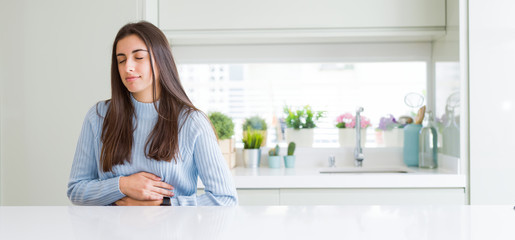 Wide angle picture of beautiful young woman sitting on white table at home with hand on stomach because nausea, painful disease feeling unwell. Ache concept.
