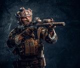 Elite unit, special forces soldier in camouflage uniform holding assault rifle and aiming with optical sight. Studio photo against a dark textured wall