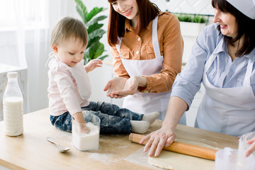 Little baby girl is sitting on the wooden table at kitchen and having fun with sugar. Grandmother and her daughters are baking cookies. Happy women in white aprons baking together. Mothers Day