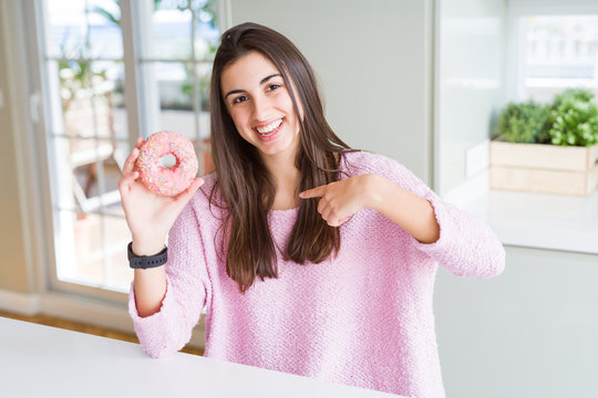 Beautiful young woman eating pink chocolate chips donut with surprise face pointing finger to himself