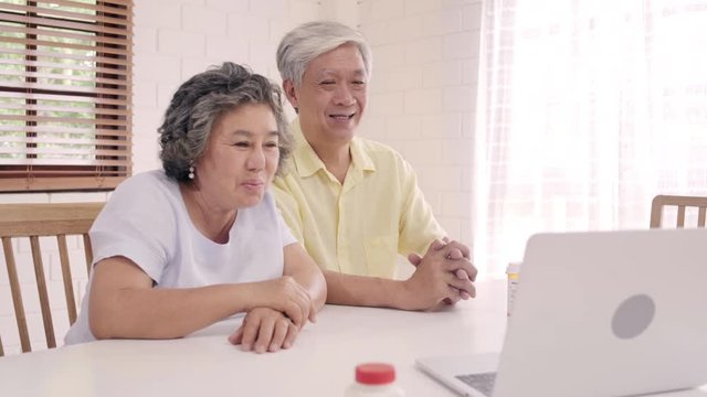 Asian elderly couple using laptop conference with doctor about medicine information in living room, couple using time together while lying on sofa at home. Senior family health at home concept.