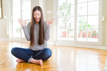 Beautiful Asian woman sitting barefoot on the floor at home celebrating mad and crazy for success with arms raised and closed eyes screaming excited. Winner concept