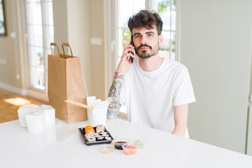 Young man eating asian sushi from home delivery and ordering food talking on smartphone with a confident expression on smart face thinking serious