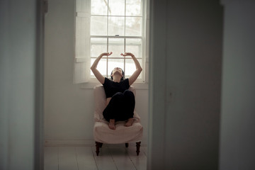 Fototapeta premium Thoughtful woman with arms raised looking away while relaxing by window on chair seen through doorway at home