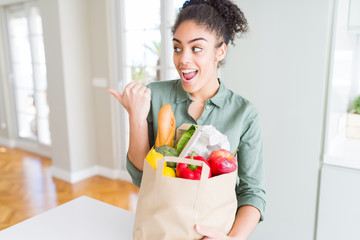 Young african american girl holding paper bag of groceries from supermarket pointing and showing with thumb up to the side with happy face smiling