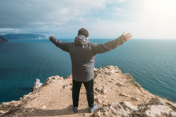 Man traveller stands on top of rock cliff with arms outstretched and looking at sea landscape, freedom of adventure concept