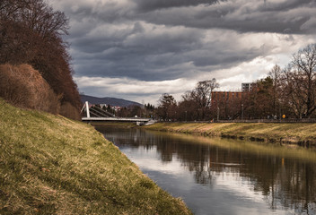 Fototapeta na wymiar Image - City in Slovakia (Nitra) with Nitra river with bridge on background - stormy weather. Photo of park in the middle of European city. Dark landscape photo in spring before a storm.