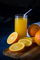 A glass of Fresh Orange Juice with a Tube, A Wooden Cooking Table, Orange slices, Oranges, white Cloth on a Dark Blue Background