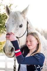 happy blonde teenager girl holding white/grey handsome horse. Sun is back lighting the scene and both are looking toward camera.
