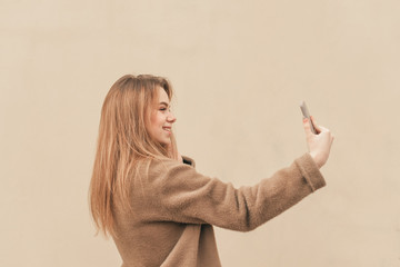 Sweet girl in a stylish spring dress, wearing a coat, standing on the background of a beige wall and takes selfie on a smartphone. Fashionable girl takes her photo on the street, background beige.
